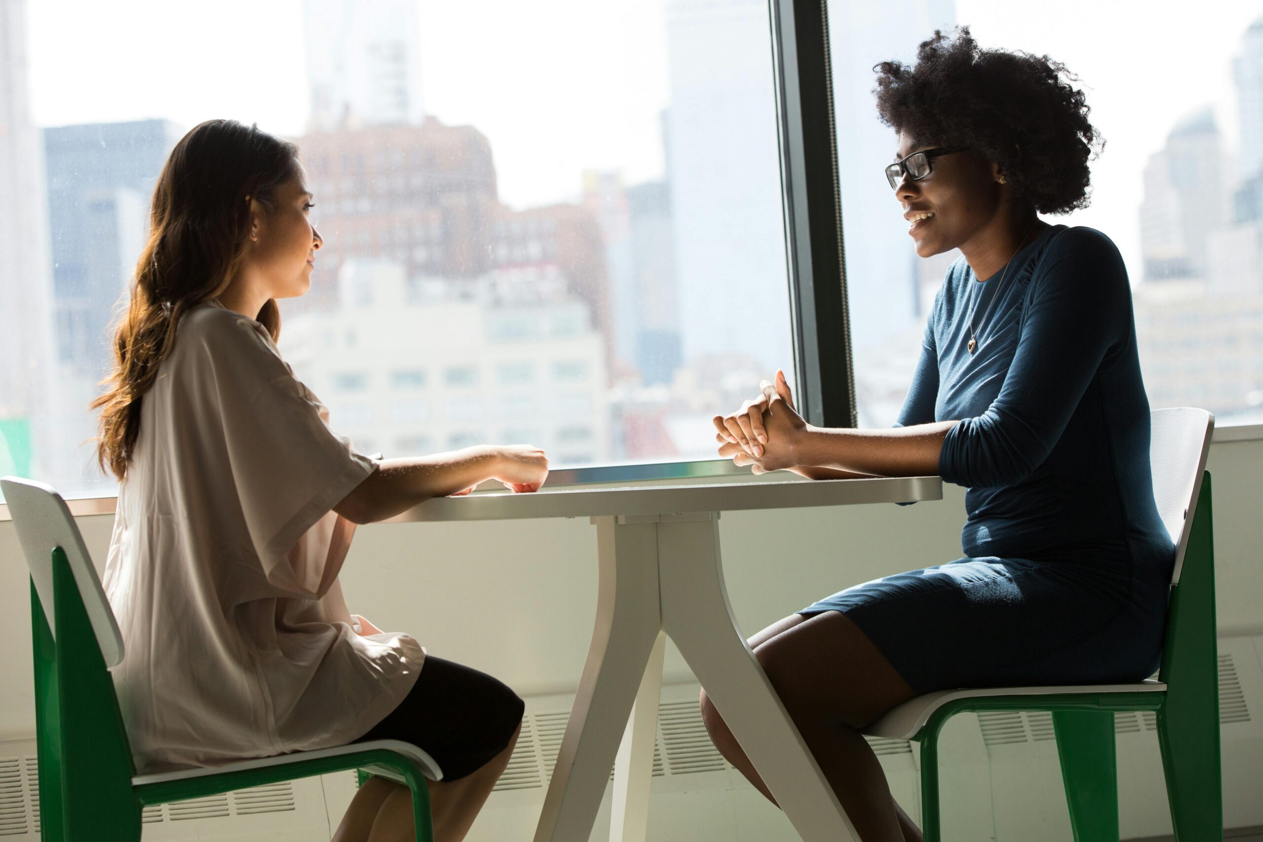 Two women sitting and talking at a table with a city view from the window. Digital Transformation